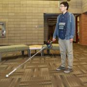A man standing in a building lobby with his arm outstretched holding a cane. The can has a white roller ball tip and a box and wires near the handle. 